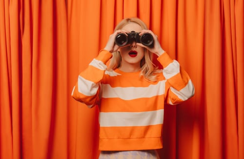 Frau mit blonden Haaren vor einem orangenen Vorhang schaut durch ein Fernglas
