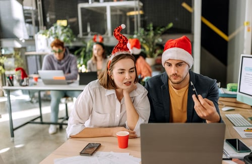 Ein Mann und eine Frau mit Weihnachtsmannmützen schauen in einem Büro gemeinsam auf einen Laptop-Bildschirm.