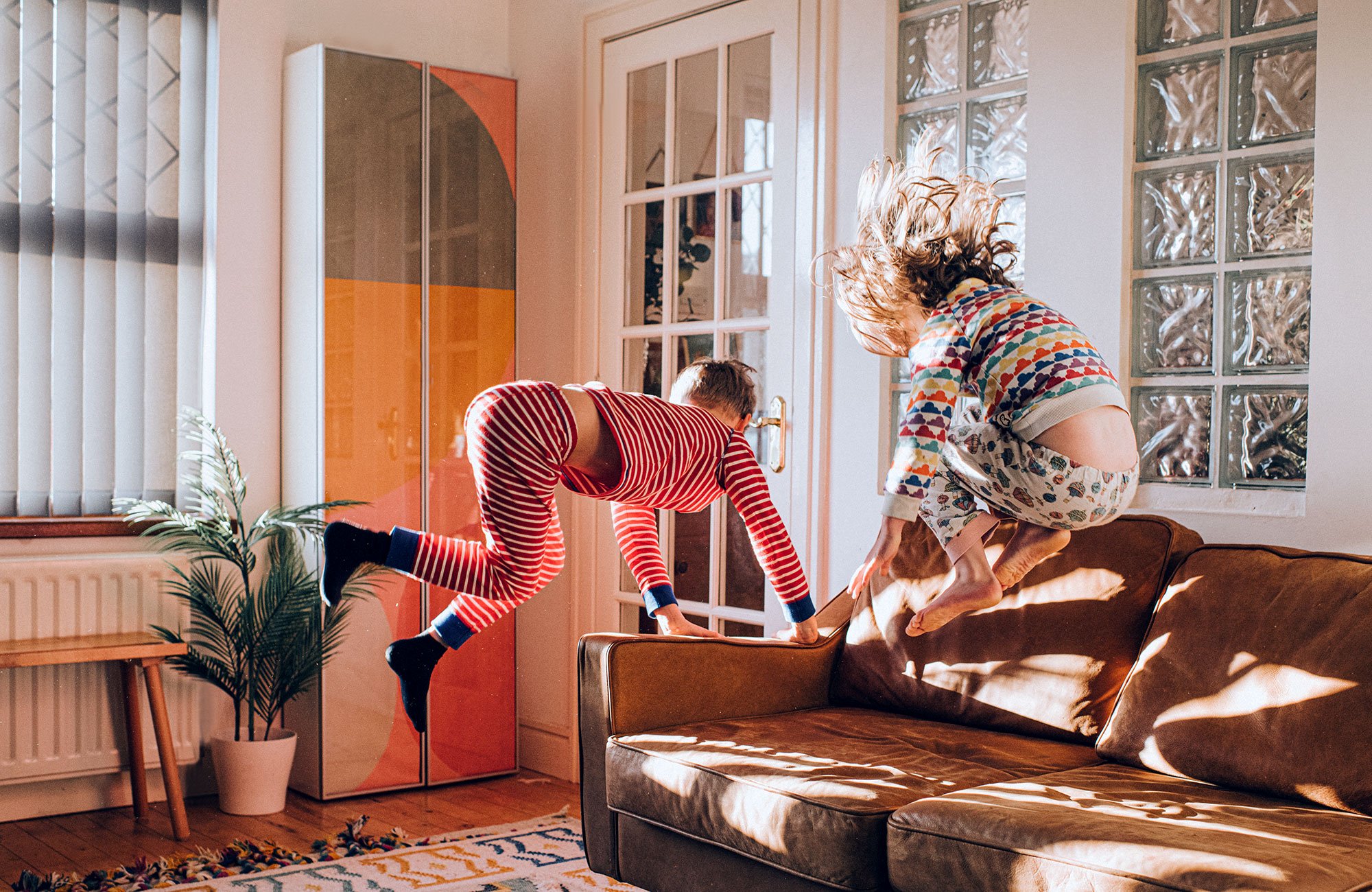 Two school aged white children wearing Christmas pajamas bounce on a brown leather couch in a sunny living room. 