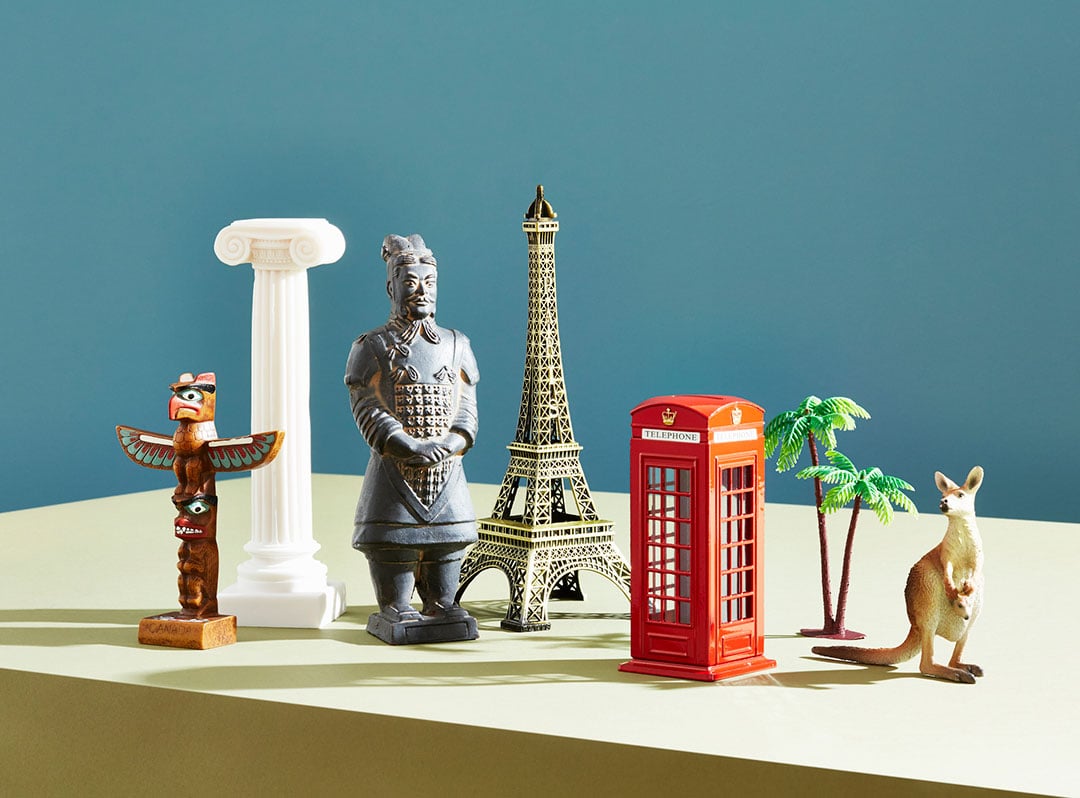 A group of small travel-themed figurines sitting on a beige table in front of a blue background. The group includes a wooden totem, a white column, a stone Chinese soldier, the Eiffel Tower, a Red telephone box, plastic palm trees, and a kangaroo.